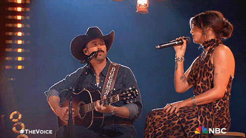 A Man In A Cowboy Hat Sings Into A Microphone While A Woman Sings Behind Him GIF