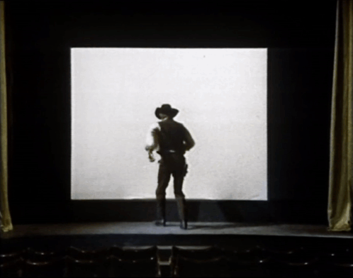 A Man In A Cowboy Hat Stands In Front Of A Large Screen GIF