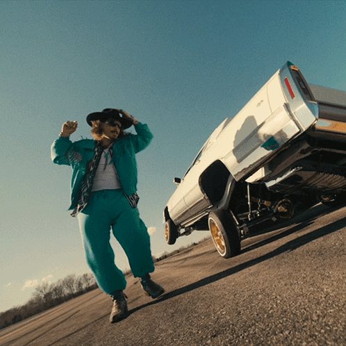 A Man In A Cowboy Hat Stands Next To A White Car On Its Rear Wheels GIF
