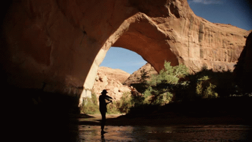 A Man In A Cowboy Hat Stands Under A Large Rock Archway GIF