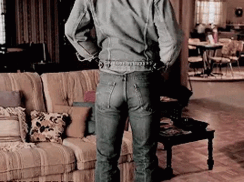 A Man In A Denim Jacket And Jeans Is Standing In A Living Room With His Hands Behind His Back . GIF