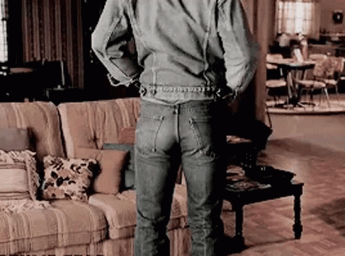 A Man In A Denim Jacket And Jeans Is Standing In A Living Room With His Hands Behind His Back . GIF