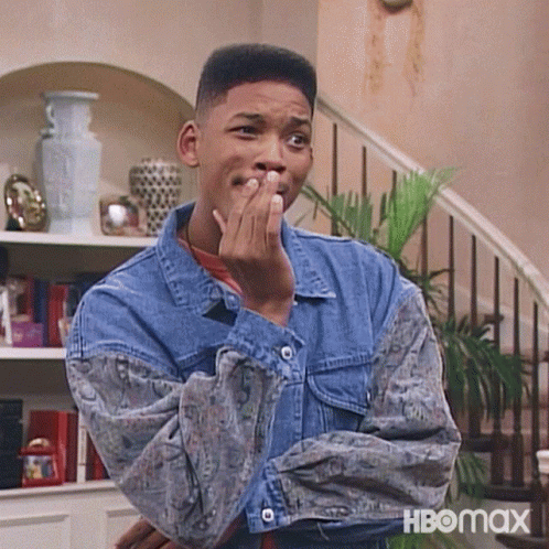 A Man In A Denim Jacket Is Covering His Mouth With His Hand While Standing In Front Of A Staircase . GIF
