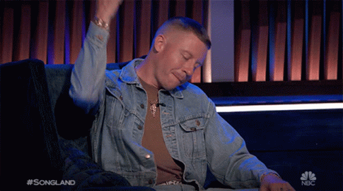 A Man In A Denim Jacket Is Sitting On A Couch With His Arms In The Air And The Words Songland On The Bottom Right GIF