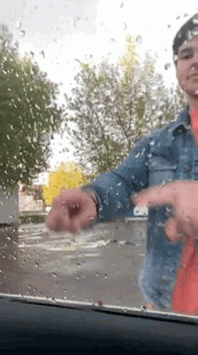 A Man In A Denim Jacket Is Standing In Front Of A Car Windshield With Rain Drops On It . GIF