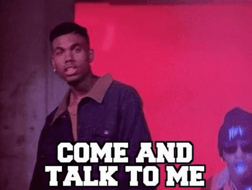 A Man In A Denim Jacket Is Standing In Front Of A Red Wall And Talking To Another Man . GIF