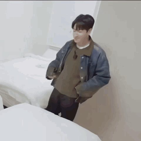 a man in a denim jacket is standing next to a bed in a room