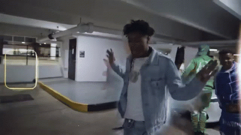 A Man In A Denim Jacket Stands In A Parking Garage With His Arms Outstretched GIF