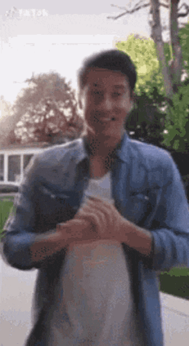A Man In A Denim Shirt And White Shirt Is Standing In Front Of A Tree . GIF