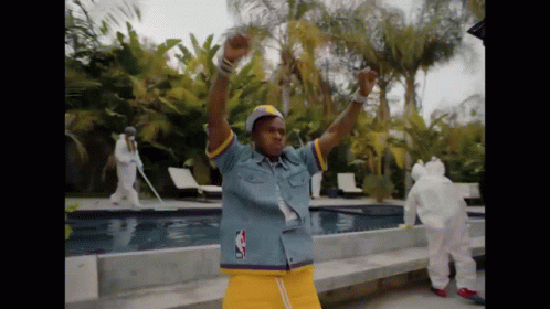 A Man In A Denim Shirt And Yellow Shorts Is Dancing In Front Of A Swimming Pool GIF