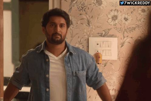 A Man In A Denim Shirt Is Standing In Front Of A Wall With Flowers On It And The Words 7Wickreddy Below Him GIF