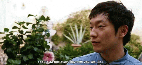 A Man In A Denim Shirt Is Talking To Another Man In Front Of A Garden . GIF