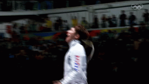 A Man In A Fencing Uniform Is Dancing In Front Of A Crowd With The Olympics Logo In The Background GIF
