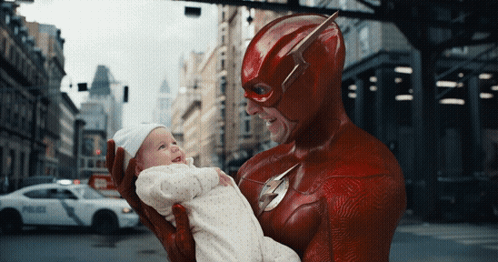 A Man In A Flash Costume Is Holding A Baby In His Arms GIF