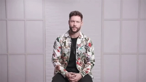 A Man In A Floral Jacket Is Sitting In Front Of A Wall With The Words Love Conquers All Above Him GIF