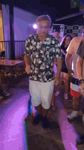 A Man In A Floral Shirt And White Shorts Is Dancing In A Crowd Of People At A Party . GIF