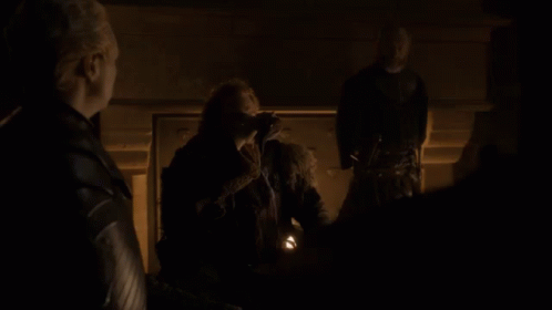 A Man In A Fur Coat Stands In A Dark Room With Two Other People GIF