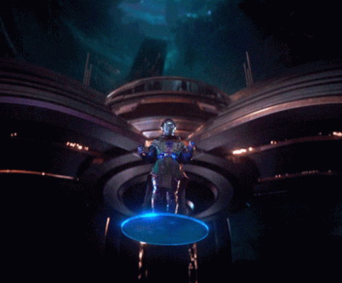 A Man In A Futuristic Outfit Is Standing In A Dark Room With A Blue Light Behind Him GIF