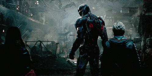 A Man In A Futuristic Suit Stands In A Dark Room With Two Other People GIF
