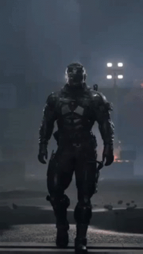 A Man In A Futuristic Suit With A Helmet On Stands In The Dark GIF