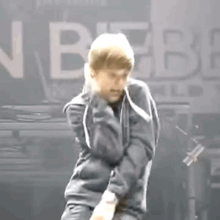 A Man In A Gray Jacket Is Dancing On A Stage In Front Of A Sign That Says Justin Bieber . GIF