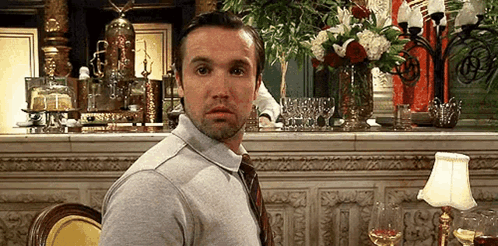 A Man In A Gray Shirt And Tie Is Sitting At A Table In A Restaurant With A Vase Of Flowers In The Background . GIF
