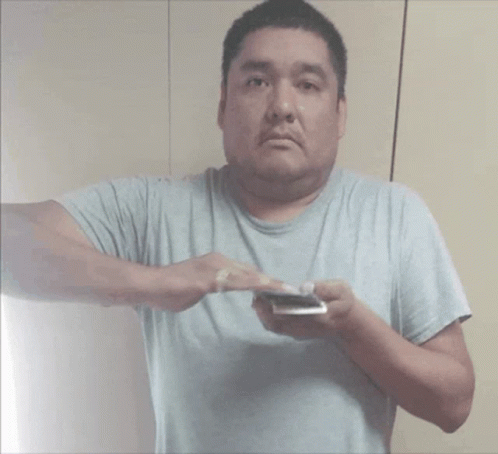 A Man In A Gray Shirt Is Holding A Cell Phone GIF
