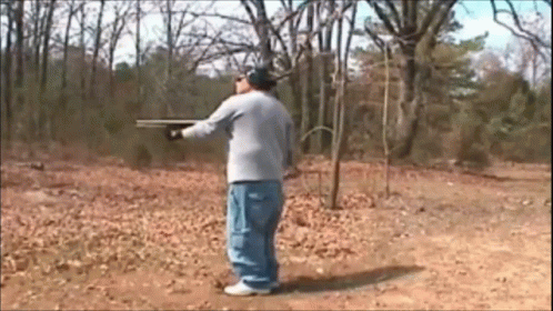 A Man In A Gray Shirt Is Holding A Shotgun In A Field GIF