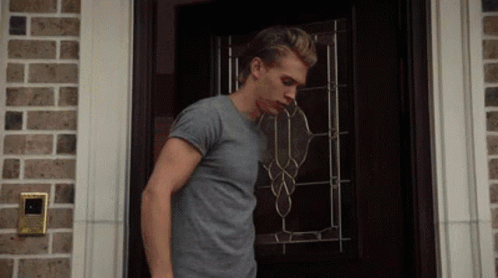 A Man In A Gray Shirt Is Standing In Front Of A Door GIF