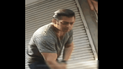 A Man In A Gray Shirt Is Standing In Front Of A Shutter GIF