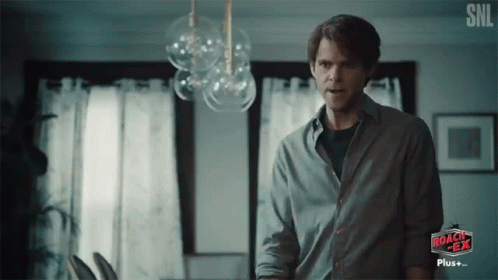 A Man In A Gray Shirt Is Standing In Front Of A Sign That Says Roach Tea GIF