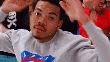 A Man In A Gray Sweatshirt Is Making A Funny Face While Sitting In A Crowd Of People . GIF