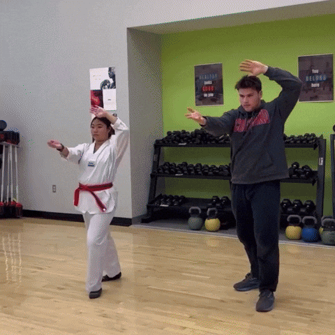 A Man In A Gray Sweatshirt Stands Next To A Woman In A Karate Uniform GIF