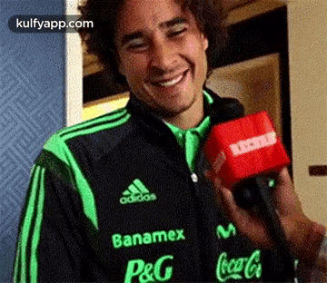 A Man In A Green And Black Adidas Jacket Is Holding A Red Microphone And Smiling . GIF