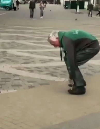 A Man In A Green And Black Jacket Is Tying His Shoes On A Sidewalk GIF