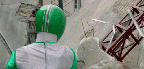 A Man In A Green And White Costume Is Standing In Front Of A Destroyed Building GIF