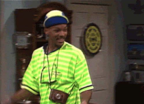A Man In A Green And Yellow Striped Shirt Is Standing In Front Of A Wall With A Dart Board On It . GIF