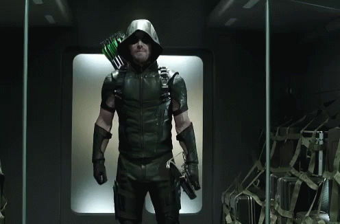 A Man In A Green Arrow Costume Is Standing In A Dark Room Holding A Bow And Arrow . GIF