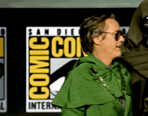 A Man In A Green Cape Is Standing In Front Of A Comic Con Logo GIF