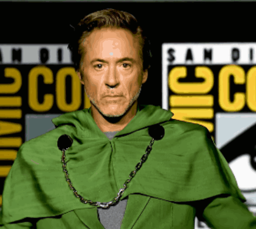 A Man In A Green Cape Stands In Front Of A Comic Con Sign GIF