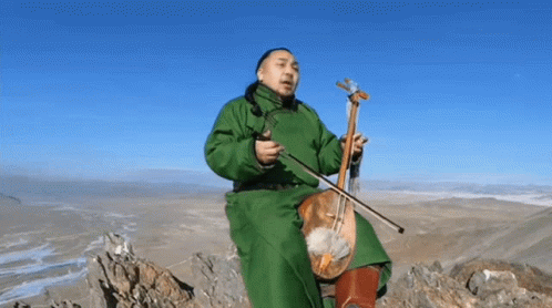 A Man In A Green Coat Is Sitting On Top Of A Mountain Playing A Violin . GIF