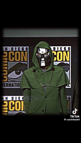 A Man In A Green Hooded Jacket Stands In Front Of A Comic Con Poster GIF