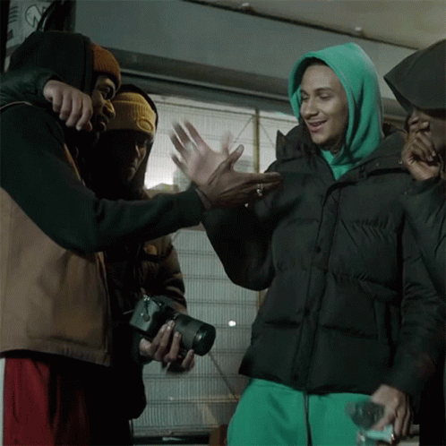 A Man In A Green Hoodie Holds A Camera And Talks To Another Man In A Black Jacket GIF