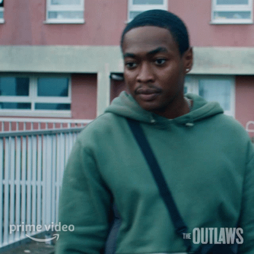 A Man In A Green Hoodie Is Standing In Front Of A Building With The Outlaws Written On It GIF