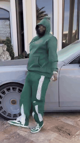 A Man In A Green Hoodie Is Standing In Front Of A Silver Car GIF