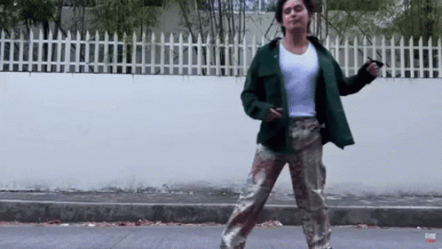 A Man In A Green Jacket And Camo Pants Is Dancing On A Street GIF