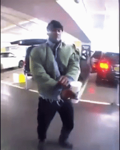 A Man In A Green Jacket Is Holding A Cup Of Coffee In A Parking Garage GIF