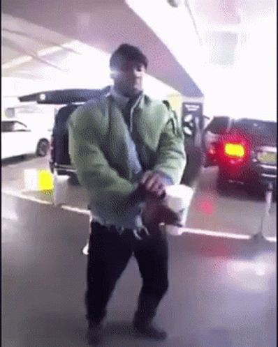 A Man In A Green Jacket Is Holding A Cup Of Coffee In A Parking Garage GIF