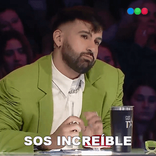 A Man In A Green Jacket Is Sitting At A Table With A Cup That Says Sos Increible On It GIF