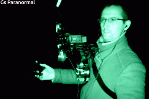 A Man In A Green Jacket Is Standing In A Dark Room With Gs Paranormal Written On The Bottom Right GIF